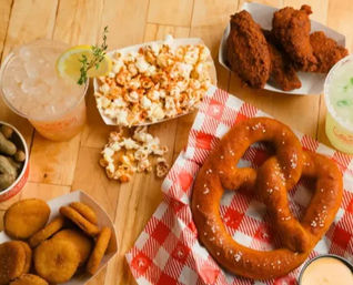 Overhead of a casual American snack spread on a wooden table: large salted soft pretzel on red-check paper, popcorn, crispy fried chicken wings, fried pickle chips, a cup of boiled peanuts, dipping sauce and two iced lemon drinks.