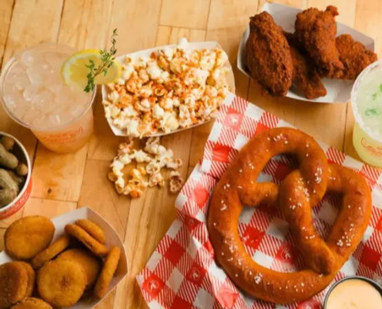 Overhead of a casual American snack spread on a wooden table: large salted soft pretzel on red-check paper, popcorn, crispy fried chicken wings, fried pickle chips, a cup of boiled peanuts, dipping sauce and two iced lemon drinks.