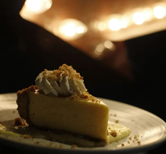 Creamy slice of cheesecake topped with whipped cream and graham-cracker crumbs on a plate, photographed under warm bokeh restaurant lights for a cozy dessert presentation.