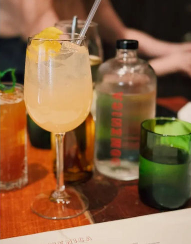 Pale peach cocktail with ice and lemon twist in a wine glass on a wooden table, next to a clear water bottle and green tumbler at a cozy bar or restaurant