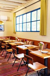 Cheerful sunlit neighborhood cafe interior with blue-framed windows, yellow curtains, wooden tables, striped cushions, small vases of flowers and a colorful striped rug.