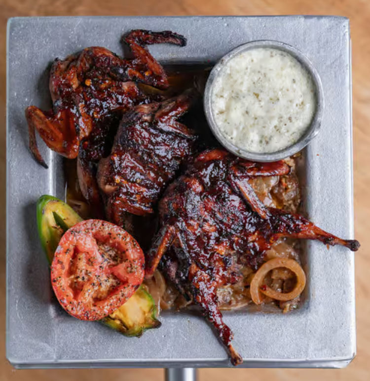 Smoky glazed grilled quail on a metal tray with caramelized onions, grilled tomato and avocado slices, served with a creamy herb dipping sauce — BBQ-style plated dish.