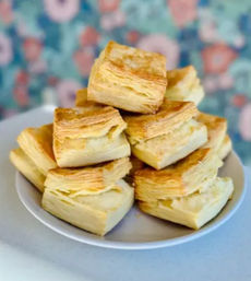 Stack of golden, flaky square biscuits with layered puff-pastry texture on a white plate, buttery breakfast pastry against a colorful floral backdrop