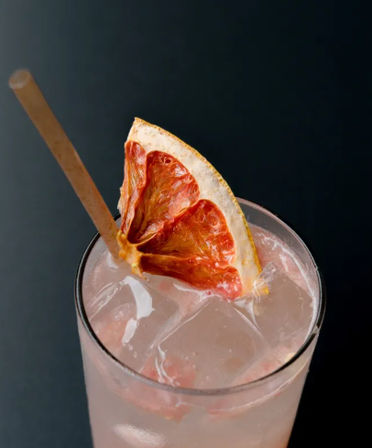Pink grapefruit cocktail in an ice-filled highball glass with a wooden straw and dehydrated grapefruit wedge garnish against a dark background