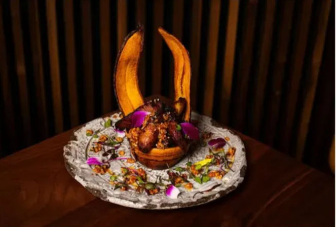 Gourmet appetizer on a rustic white plate: bite-sized fried pieces in a small bowl topped with two tall crisp plantain chips, garnished with purple edible flower petals, microgreens and crunchy bits on a dark wood table.