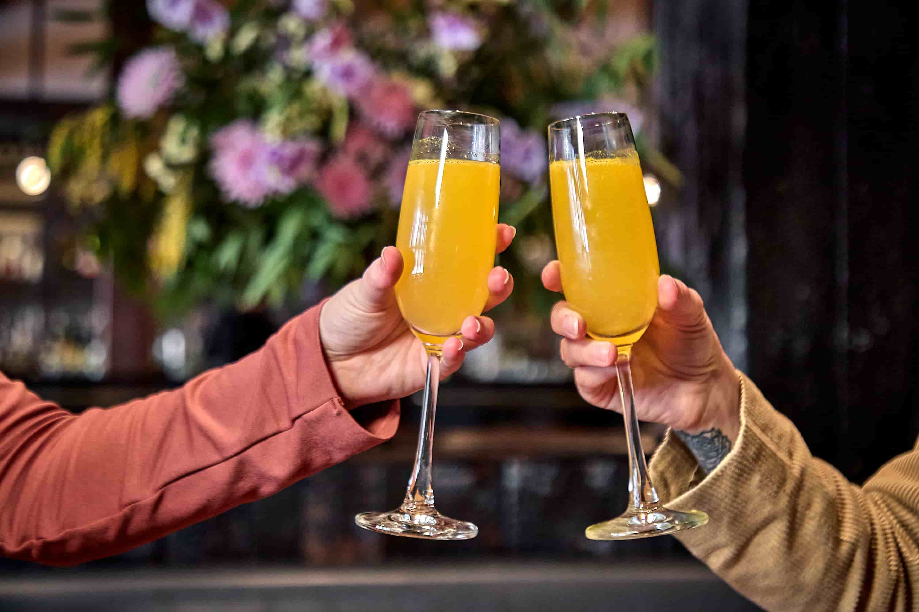 Two hands clinking champagne flutes filled with bright orange mimosas during a cozy brunch in a restaurant with blurred floral decor in the background.