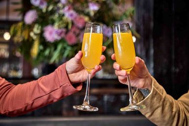 Two hands clinking champagne flutes filled with bright orange mimosas during a cozy brunch in a restaurant with blurred floral decor in the background.