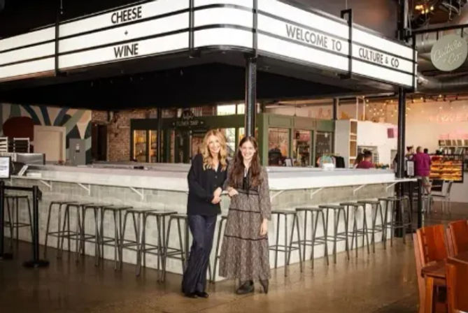 Two smiling women at a white bar in an indoor food hall beneath a lit 'CHEESE'/'WINE' marquee, surrounded by metal stools and industrial-chic decor.