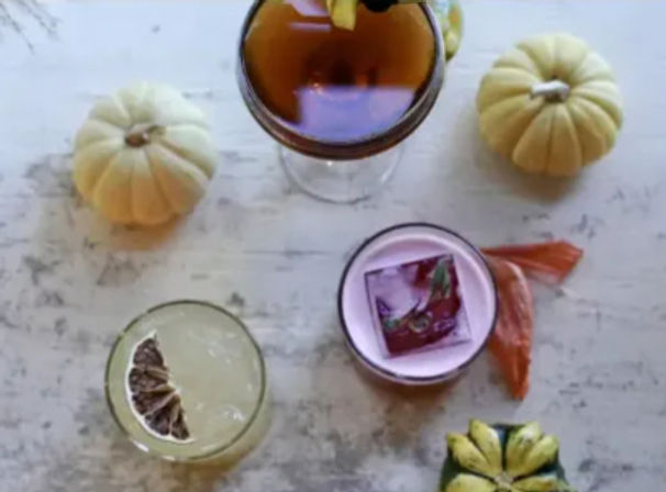 Overhead flatlay of three fall cocktails on a distressed white wooden table, surrounded by mini white pumpkins, a dried citrus slice and colorful seasonal garnishes.