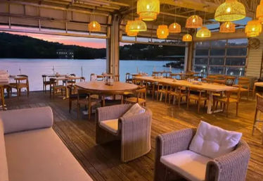 Cozy open-air waterfront dining deck at sunset with wooden tables, wicker lounge chairs and glowing rattan pendant lights overlooking a calm lake