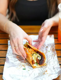 Hands holding a foil-wrapped street taco filled with charred pork, pineapple, diced onion, cilantro and sauce on a wooden table — casual outdoor dining, ready to eat.