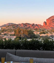 Sunset-lit red rock mountains overlooking a suburban neighborhood, seen from an outdoor patio with a striped sofa, cushions and drink glasses in the foreground.