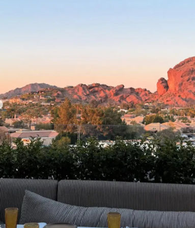 Sunset-lit red rock mountains overlooking a suburban neighborhood, seen from an outdoor patio with a striped sofa, cushions and drink glasses in the foreground.