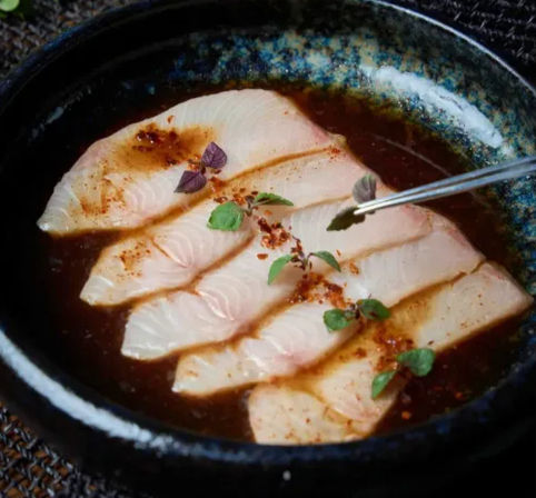 Japanese-style sashimi of thin white fish slices in a dark soy-based sauce in a rustic black bowl, sprinkled with chili flakes and micro herb leaves