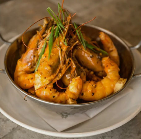 Metal bowl of whole prawns in a spicy garlic-butter sauce garnished with sliced scallions, served on a white plate — restaurant-style seafood dish