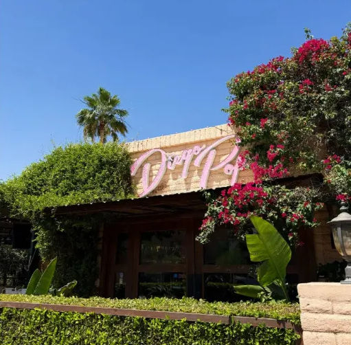 Sunlit stucco storefront with a playful pink cursive sign peeking through fuchsia bougainvillea and trimmed hedges, palm tree under a clear blue sky.