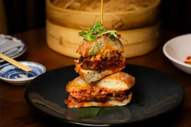Stacked Asian-fusion sandwich on a black plate with spicy pickled-vegetable filling and microgreens on top, chopsticks and a bamboo steamer in the background on a wooden table