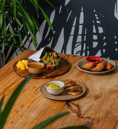 Tropical‑themed café wooden table with a colorful vegetable wrap on a wooden board, fried pastry turnovers with red dipping sauce, and grilled plantain slices with a creamy dip.