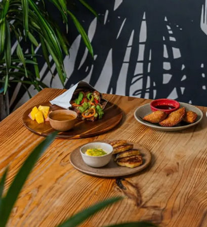 Tropical‑themed café wooden table with a colorful vegetable wrap on a wooden board, fried pastry turnovers with red dipping sauce, and grilled plantain slices with a creamy dip.