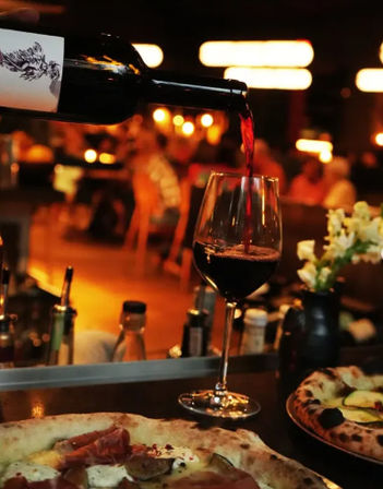 Red wine poured into a glass at a cozy dimly lit restaurant wine bar, with wood-fired pizza on the counter and warm bokeh lights in the background