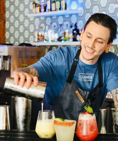 Smiling bartender pouring a shaken cocktail into a glass at a modern bar, three colorful craft cocktails garnished with lime, chili-rim and mint in the foreground and hexagon-tiled back bar with bottles.
