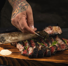 Close-up of charred bone-in ribeye sliced on a wooden board, tattooed hand spooning caviar over the steak with microgreens and seasoning piles