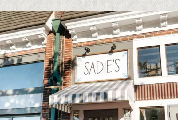 Charming downtown brick storefront with decorative white cornice, striped gray-and-white awning, wooden-framed white sign beneath two gooseneck lights, and large display windows reflecting the street.
