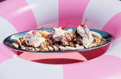 Summer-ready banana split with three ice cream scoops, whipped cream, cherries, chocolate sauce and chopped nuts on a metal dish perched on a pink-and-white striped inflatable pool float