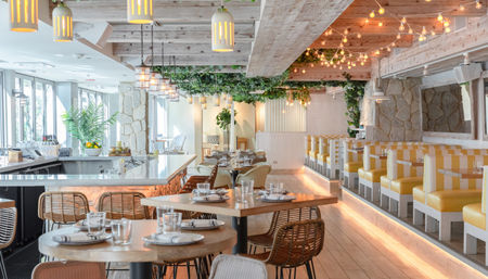 Inviting sunlit coastal restaurant interior with whitewashed wood beams, hanging pendant and string lights, marble bar, wicker chairs, and yellow-striped booth seating.