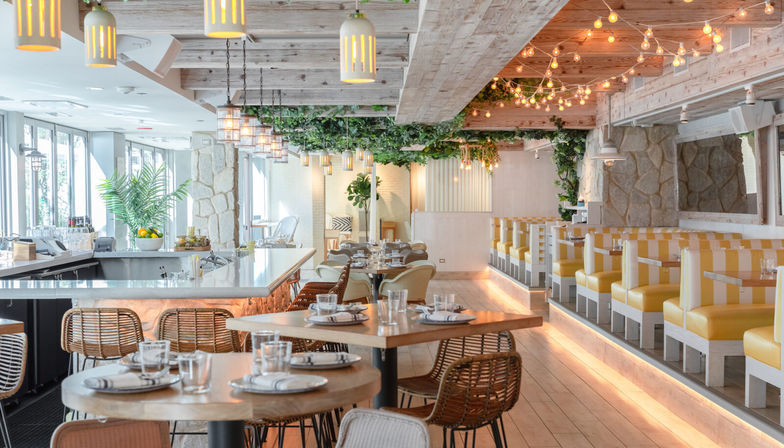 Inviting sunlit coastal restaurant interior with whitewashed wood beams, hanging pendant and string lights, marble bar, wicker chairs, and yellow-striped booth seating.