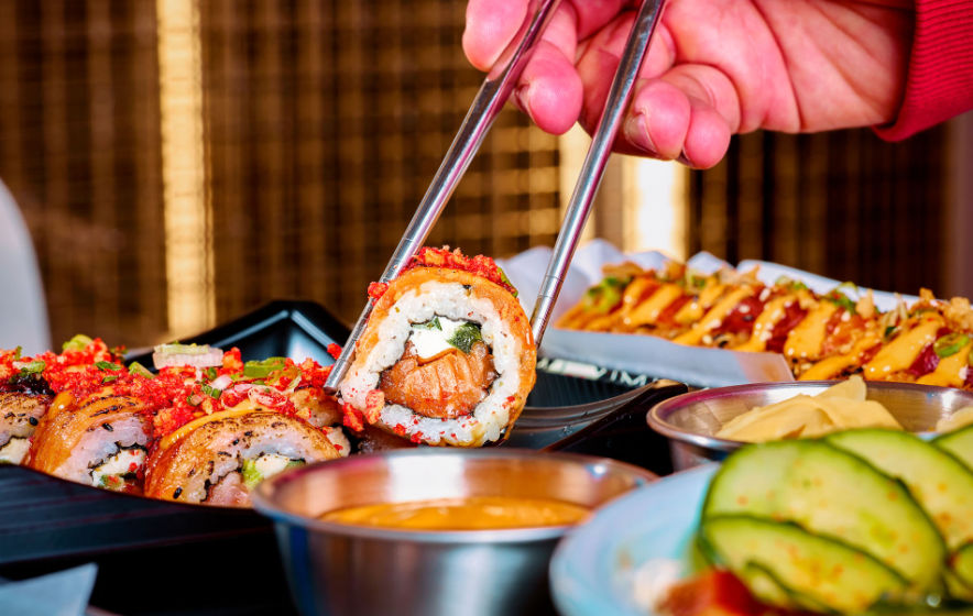 Hand with chopsticks lifting a salmon-filled sushi roll topped with crunchy red flakes, plated with dipping sauces, pickled ginger and cucumber slices