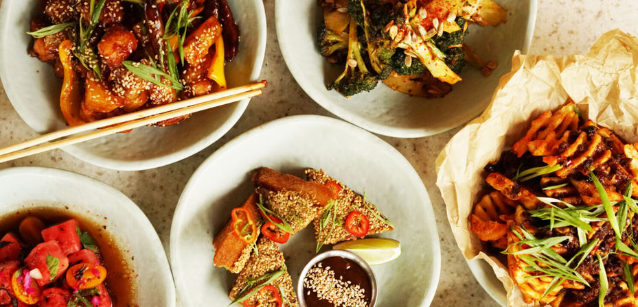Overhead of shareable Asian-inspired small plates on a light stone table: sesame-crusted fried triangles with dipping sauce and lime, sesame stir-fry with peppers and chopsticks, roasted broccoli, spicy waffle fries with scallions, and vibrant watermelon salad.