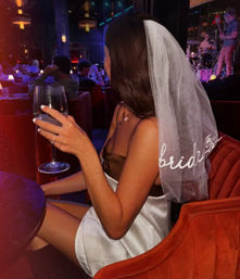 Bride-to-be wearing a veil and white satin dress holding a wine glass in a dimly lit lounge with a live band — bachelorette party nightlife