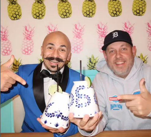 Two smiling people in a tropical-themed bar photo booth holding blue-and-white ceramic cocktail mugs with lime and straws against a pineapple wallpaper backdrop.