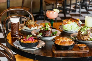 Lively urban gastropub spread on a round table: beer in a boot glass, pink margarita, skillet-baked bread, roasted meats, wedge salad with bacon and pickled onions.