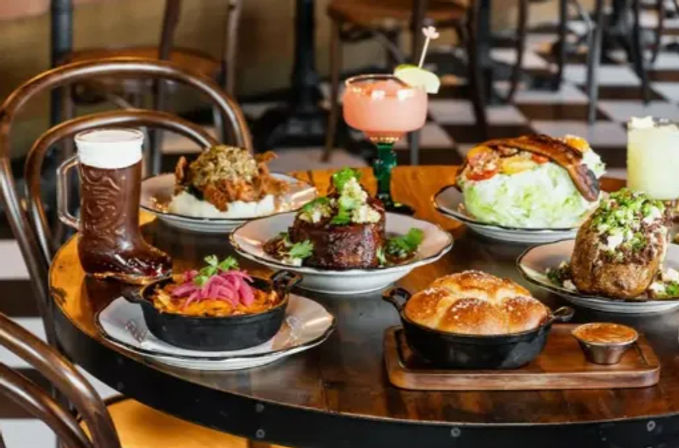 Lively urban gastropub spread on a round table: beer in a boot glass, pink margarita, skillet-baked bread, roasted meats, wedge salad with bacon and pickled onions.
