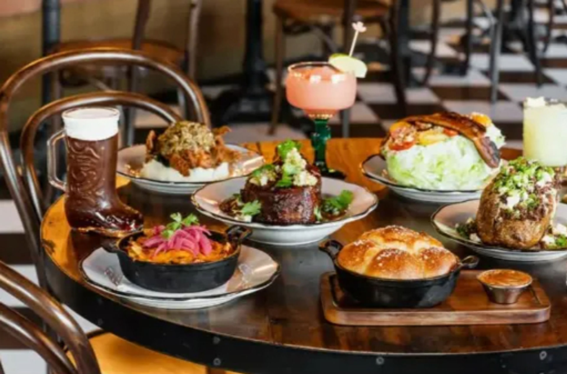 Lively urban gastropub spread on a round table: beer in a boot glass, pink margarita, skillet-baked bread, roasted meats, wedge salad with bacon and pickled onions.