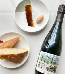 Top-down view of a sparkling wine bottle beside two white plates on a light speckled countertop — one plate with sliced baguette, the other with a caramelized sugar strip and a quenelle of sorbet, an inviting brunch-style spread.