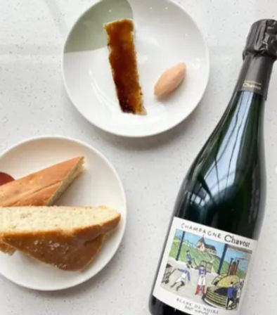 Top-down view of a sparkling wine bottle beside two white plates on a light speckled countertop — one plate with sliced baguette, the other with a caramelized sugar strip and a quenelle of sorbet, an inviting brunch-style spread.