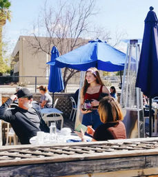 Sunny outdoor café patio with bright blue umbrellas and patio heaters; young woman in a red top holding a coffee carafe while chatting with seated diners.