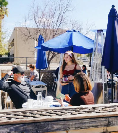 Sunny outdoor café patio with bright blue umbrellas and patio heaters; young woman in a red top holding a coffee carafe while chatting with seated diners.