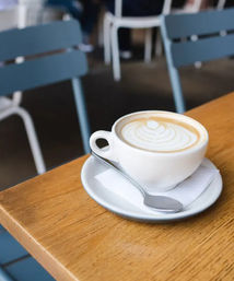 White ceramic cup of latte with leaf latte art on a saucer and spoon, sitting on a wooden cafe table with blue chairs in the background.