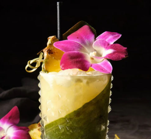 Tropical pineapple cocktail with crushed ice, pineapple wedge and bright pink orchid garnish in a textured, leaf-wrapped glass on a moody black background.