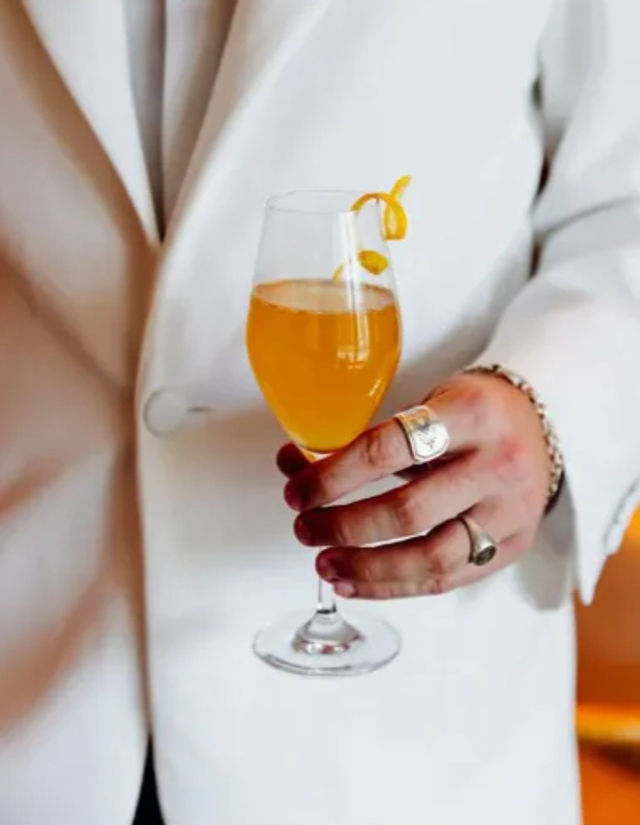 Person in a white blazer holding an amber cocktail in a stemmed wine glass garnished with a citrus twist, rings and a bracelet visible on the hand.