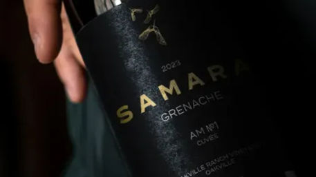 Close-up of a hand holding a matte black wine bottle labeled Grenache 2023 with gold lettering, moody dark background, elegant close-up shot