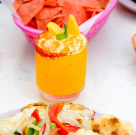 Bright mango drink in a chili-rimmed glass with ice and mango slices, served with tortilla chips and a plate of grilled vegetables on a white table