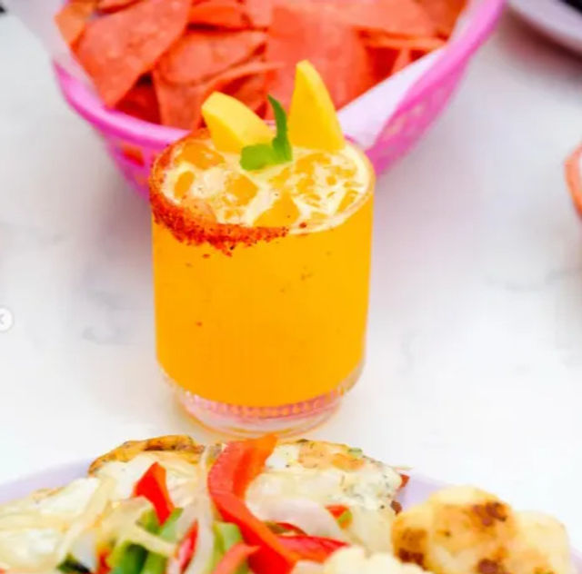 Bright mango drink in a chili-rimmed glass with ice and mango slices, served with tortilla chips and a plate of grilled vegetables on a white table