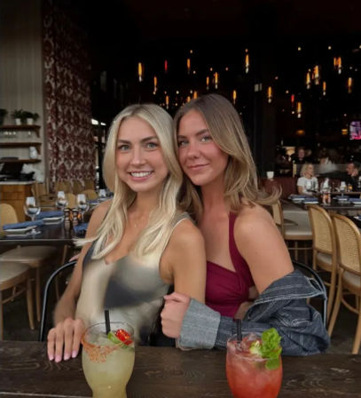 Two smiling women posing at a trendy urban restaurant table with colorful cocktails in front of them, warm pendant lights and set dining tables in the dim background.