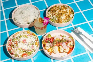 Four colorful bowls of Mexican-inspired salads and elote-style bowls topped with crumbled white cheese, radish slices, cherry tomatoes and pickled onions, centered around a lime margarita garnished with a dried lime and orchid, served on a bright blue tiled table with a fork and napkin.