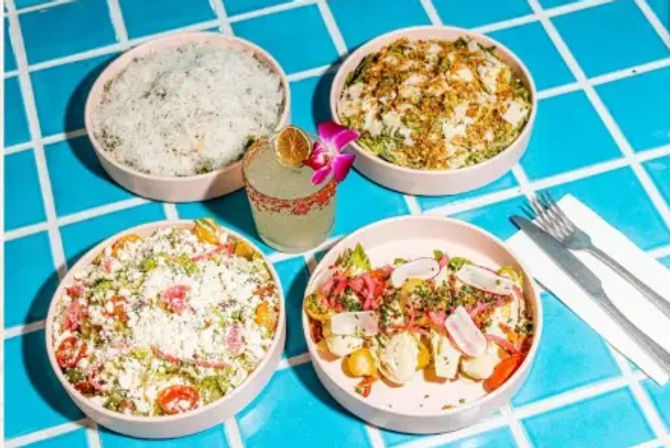 Four colorful bowls of Mexican-inspired salads and elote-style bowls topped with crumbled white cheese, radish slices, cherry tomatoes and pickled onions, centered around a lime margarita garnished with a dried lime and orchid, served on a bright blue tiled table with a fork and napkin.